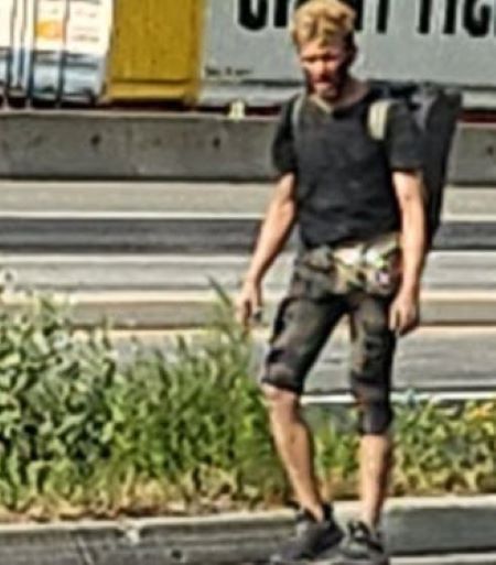 A male with blond hair, knee length shorts, a dark shirt and dark shoes, walks next to the highway. 