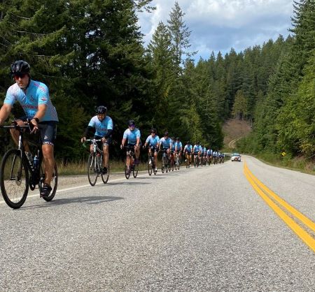 Cyclists in blue team shirts, shorts, and helmets, ride single file along the roadside. A police car with its lightbar on follows behind them.