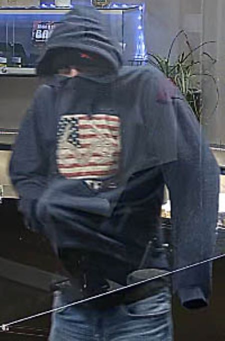 Suspect avec un capuchon : un homme porte un chandail à capuchon bleu marin avec un drapeau américain imprimé sur le devant. Le capuchon lui recouvre le nez. Il a la main dans la poche du chandail et on dirait qu’il y tient quelque chose.