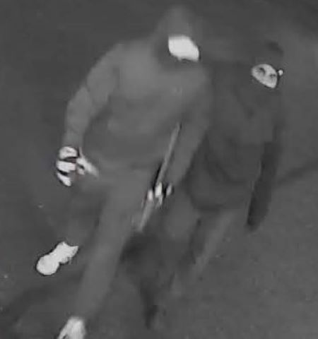 Deux suspects portant des vestes à capuchon et des masques sont filmés en train de marcher dans une vidéo de surveillance en noir et blanc. 