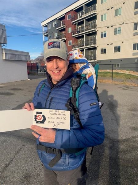 A man in a blue jacket with a baby carrier sporting a baby doll on his back, holds up a card with a message advising people not to use their phone while driving.   